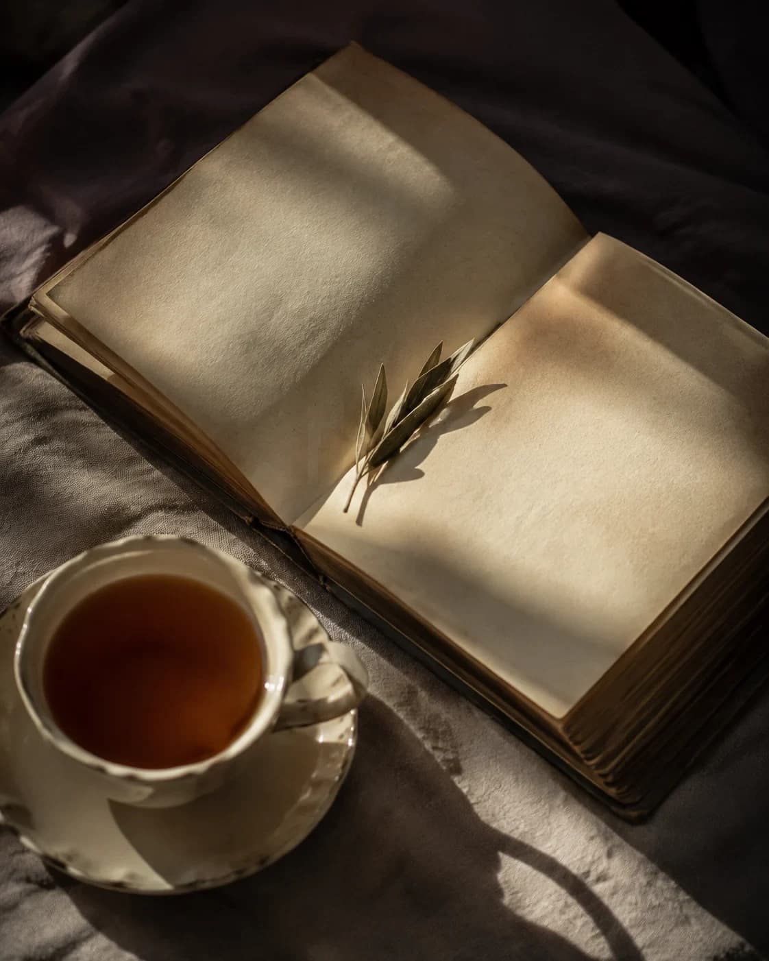 An open book with pale pages and an olive sprig beside a cream cup of tea on linen.