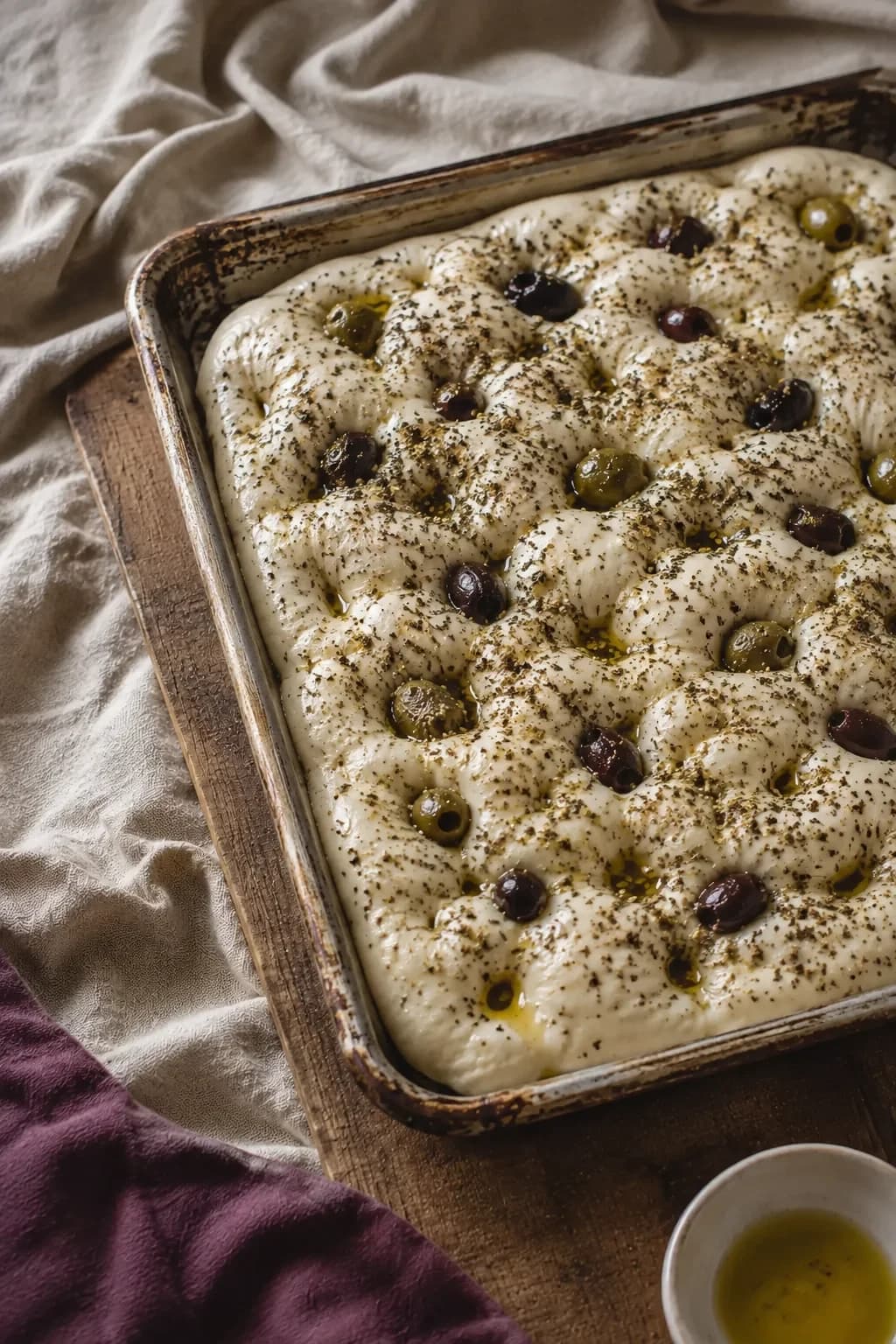 Pale dimpled focaccia dough topped with green and dark olives and zaatar in a metal tray.