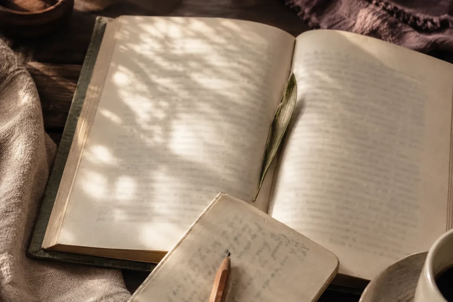 An open book with an olive leaf bookmark beside a notebook, pencil and partly visible cup.